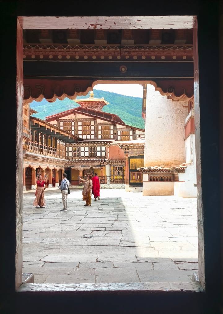 Visitors explore the ornate courtyard of a traditional Bhutanese monastery with intricate woodwork and mountain scenery beyond.