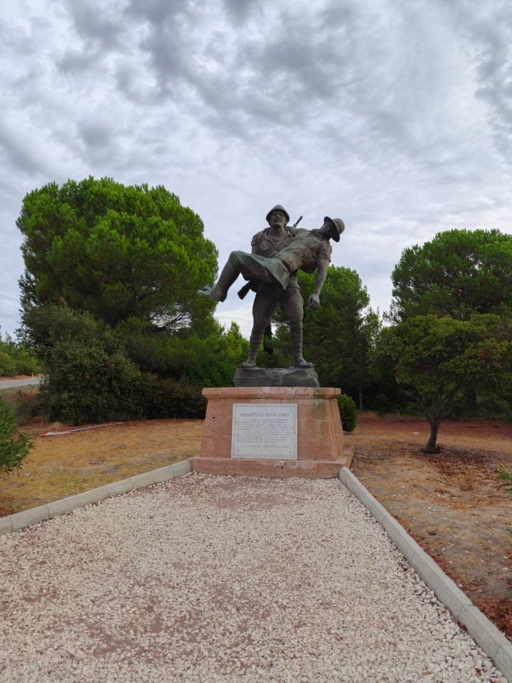 Bronze statue of a World War I Turkish soldier carrying a wounded comrade stands among pine trees.