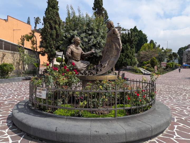 Bronze sculpture of Juan Diego and the Virgin of Guadalupe surrounded by rose bushes in a plaza