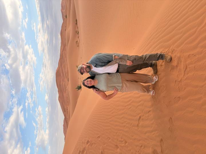Smiling couple stand arm in arm on rippled orange dunes beneath a partly cloudy sky in the Moroccan Sahara.