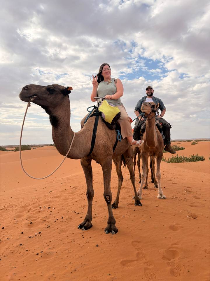 Two travelers ride camels across rolling orange sand dunes, a cloudy sky overhead.