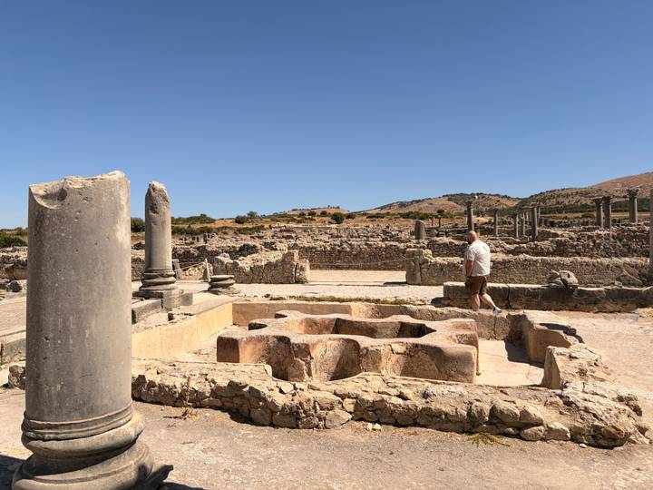 A lone visitor surveys the ruined Roman columns and mosaicked foundations of Volubilis beneath a bright blue sky.