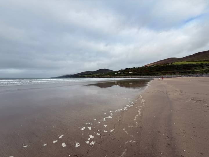 Wide, empty sandy beach with gentle surf and distant green hills beneath a grey, cloud-filled sky.