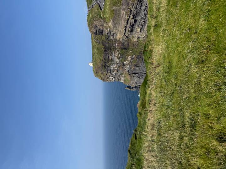 Steep grassy cliff jutting into a calm blue Atlantic with a small white building perched on top.