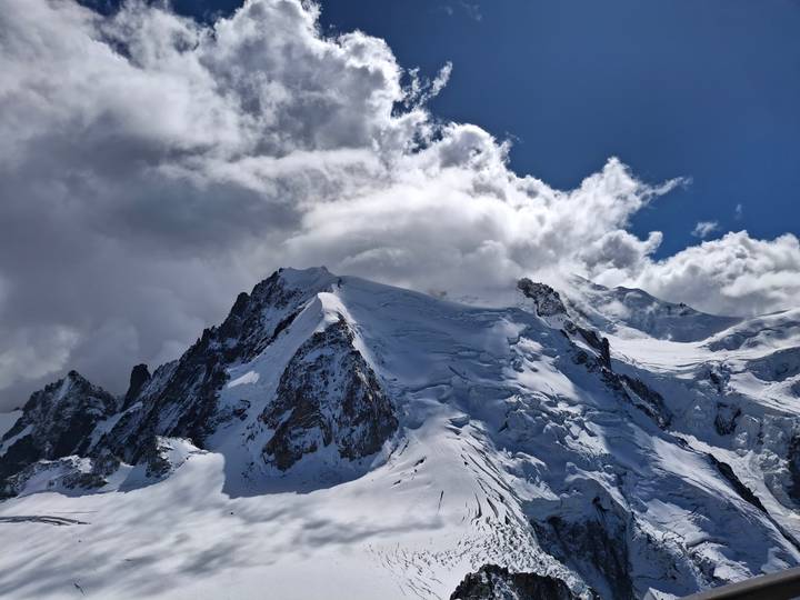 Dramatic snow-covered alpine peaks and glaciers piercing swirling clouds under a deep blue sky.