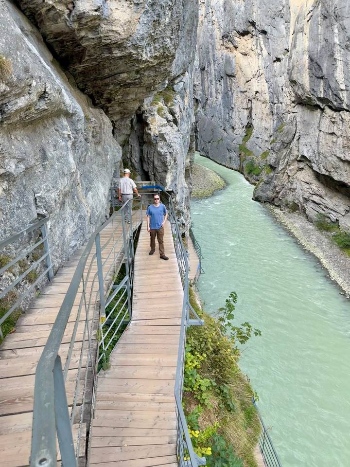 Visitors on a narrow metal walkway carved into a steep cliff above a milky turquoise river gorge.