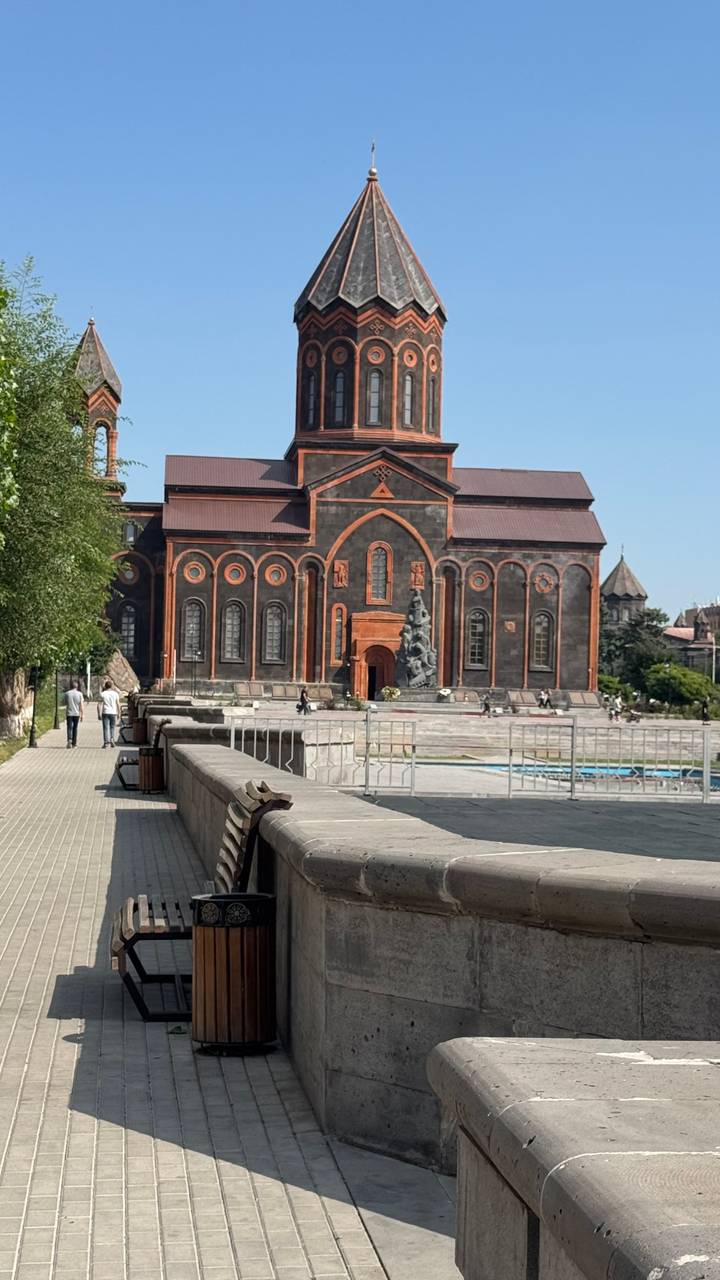 Dark-stone Armenian church with ornate orange trims dominating a spacious city square.
