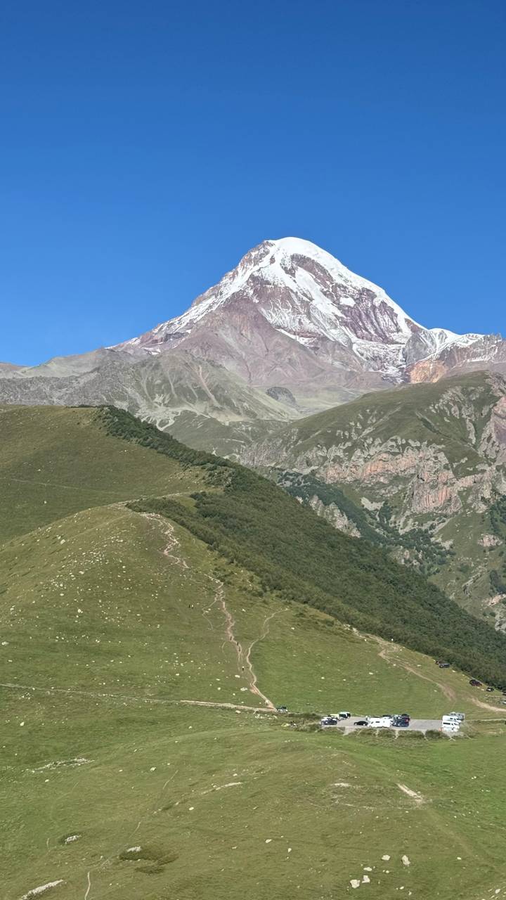 Snow-capped Mount Kazbek rising above rolling green ridges under a clear blue sky.