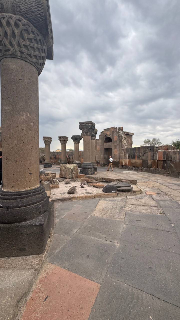 Traveler exploring the columned ruins of Zvartnots Cathedral under a cloudy sky.