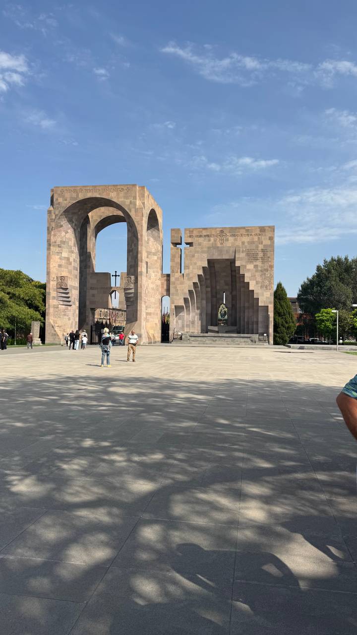 Spacious stone plaza with a monumental modern arch and cross, visitors scattered around.