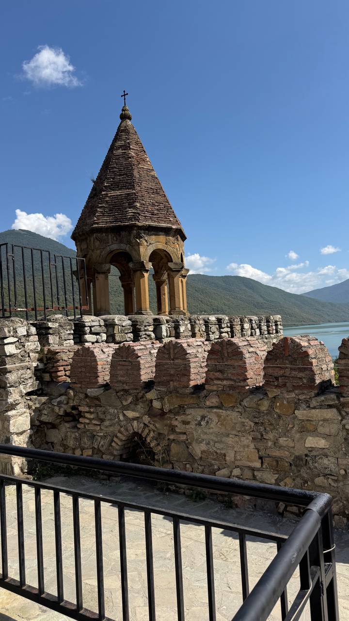 Stone bell tower of a medieval fortress overlooking a turquoise reservoir and forested mountains.