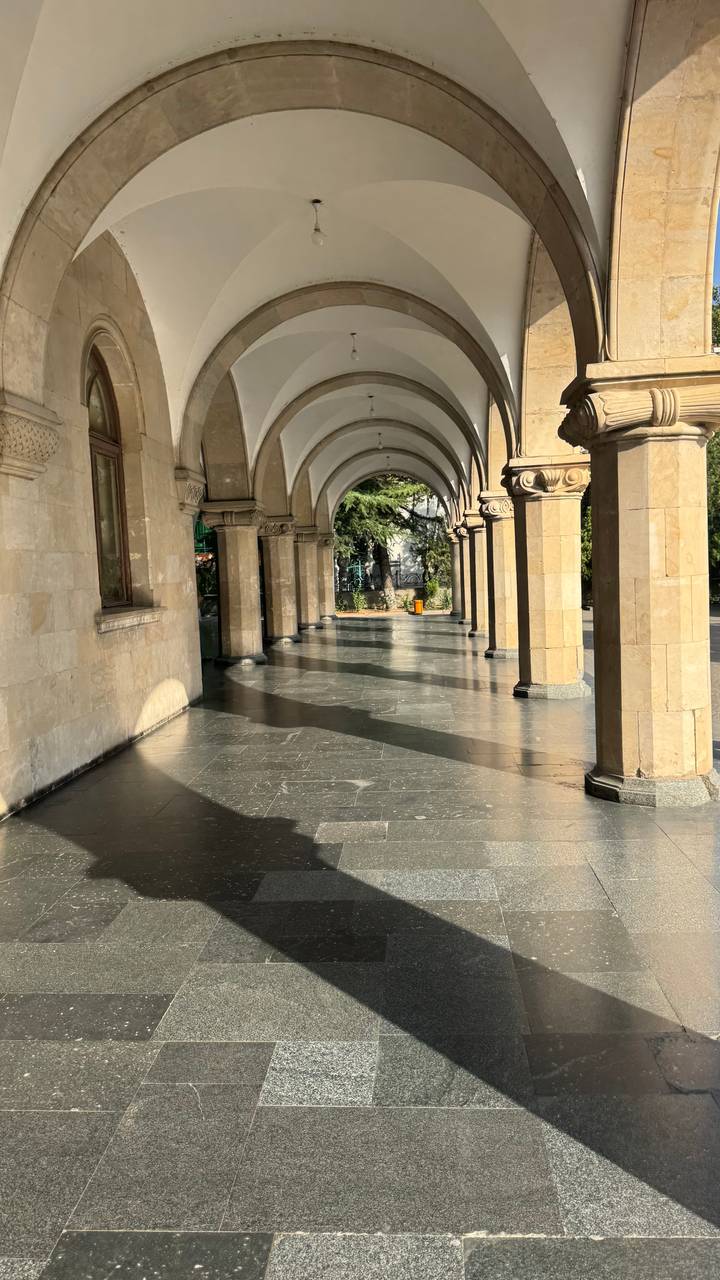 Sunlit stone arcade with long shadows on polished floor leading to a garden.