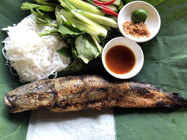 A grilled whole fish served with rice noodles, fresh herbs, cucumber and dipping sauces on a banana leaf.