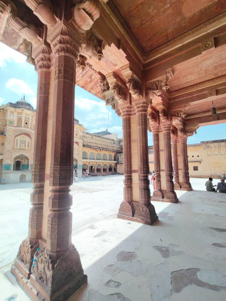 Sun-lit courtyard of Amber Fort framed by ornate sandstone pillars with tourists exploring the space.