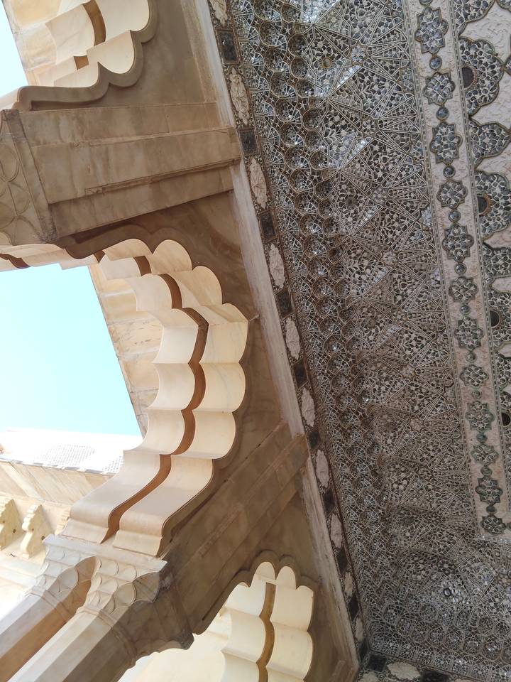 Decorative marble archway meeting a finely carved ceiling with blue sky visible through the opening.