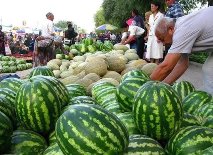 Outdoor market stall piled high with watermelons and melons, shoppers and vendors in the background.