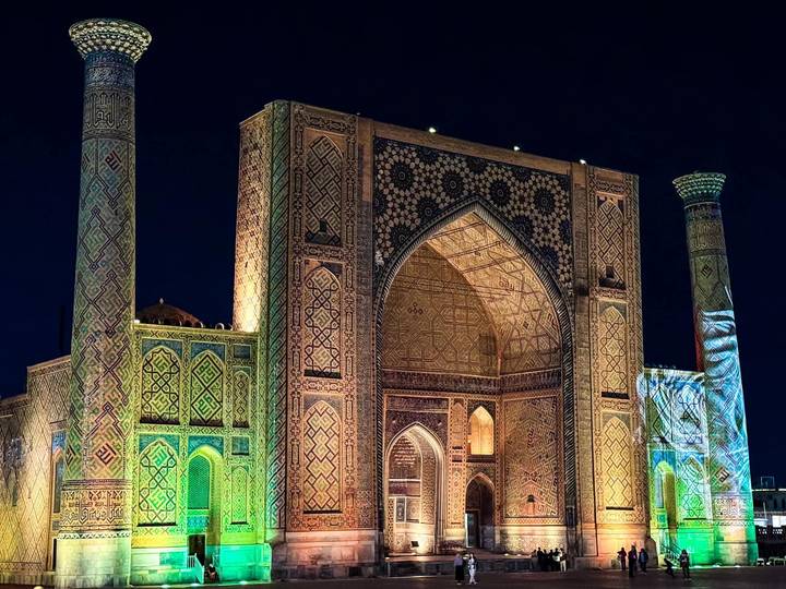 Illuminated Islamic madrasah with ornate tilework glowing against the dark night sky.