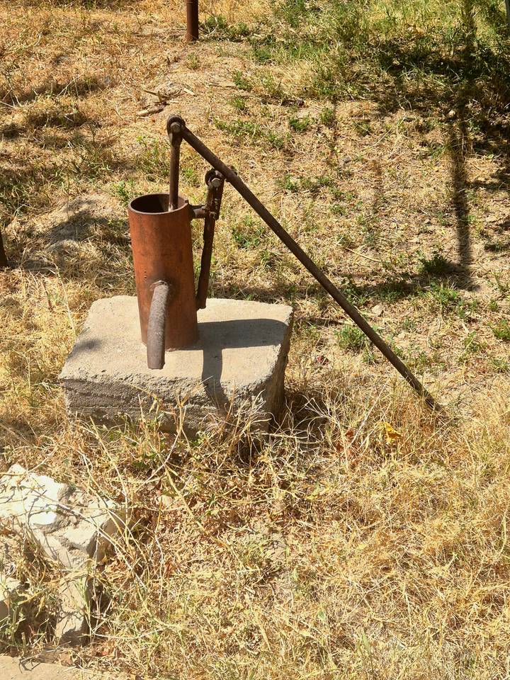 Old rusty hand water pump fixed on a concrete block in a dry grassy field.