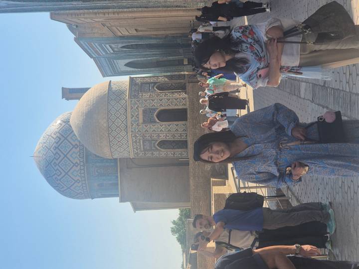 Tourists walking past the blue-tiled domes and mausoleums of Samarkand on a sunny day.
