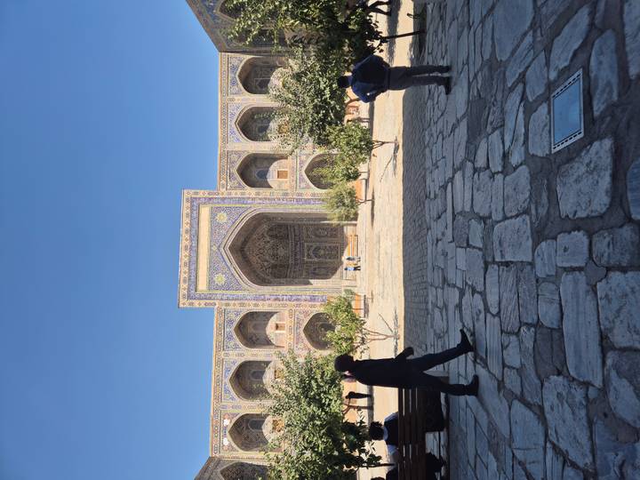 Ornate courtyard entrance of a richly tiled madrasah framed by trees and visitors in silhouette.