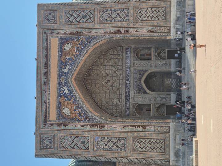 Grand facade of Sher-Dor Madrasah with intricate mosaic and visitors gathered on the steps.