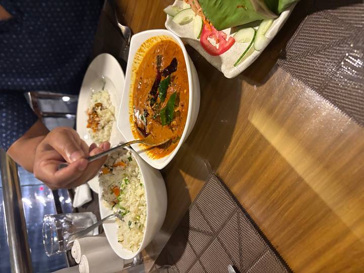 Close-up of a hand serving curry beside bowls of rice and vegetables in a restaurant.