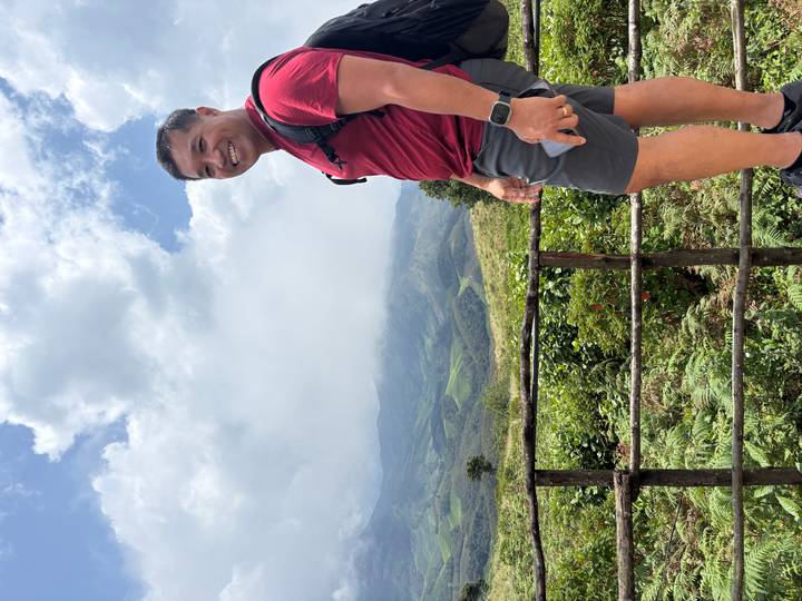 Hiker smiling at a mountain viewpoint with rolling green hills and partly cloudy sky behind.