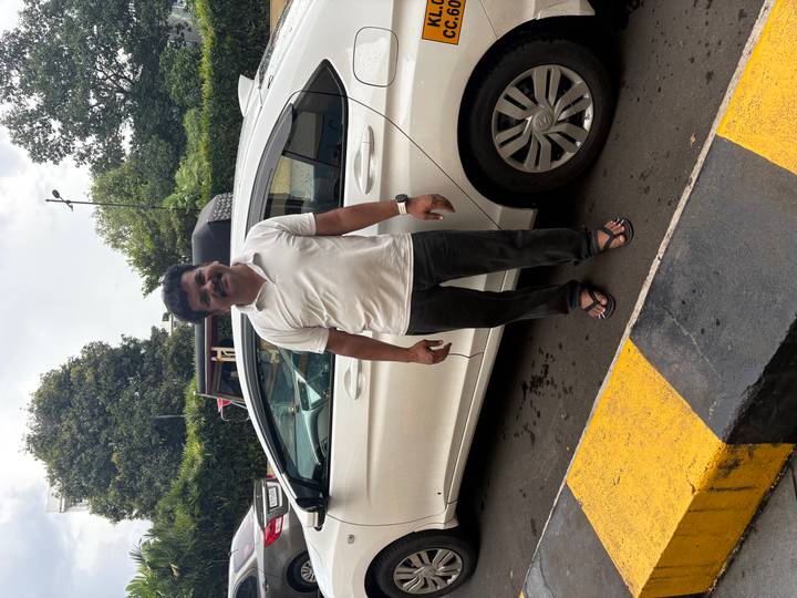 Driver standing beside a white sedan taxi in a parking area on an overcast day.