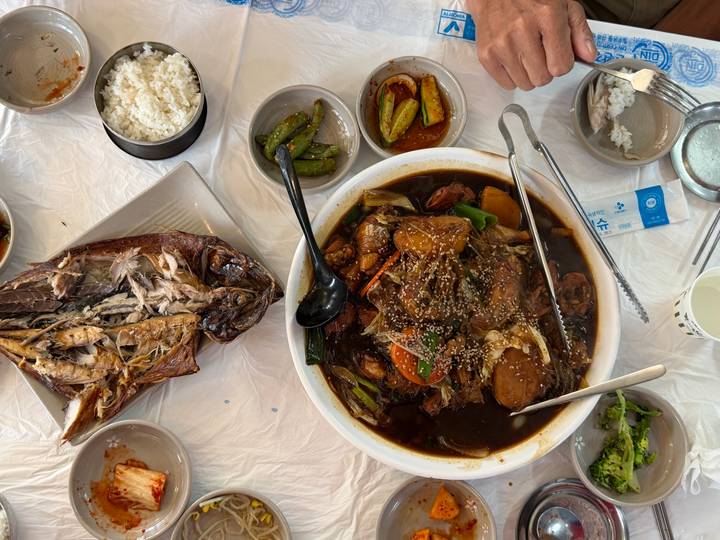 Korean seafood stew and grilled fish surrounded by assorted side dishes on a plastic-covered table.