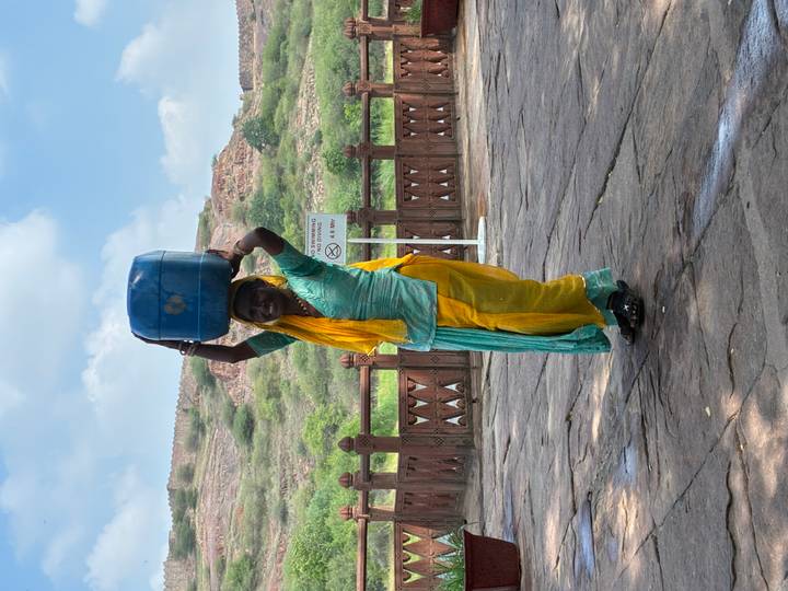 Woman in colorful traditional clothing balances a large blue container on her head against a fort backdrop.