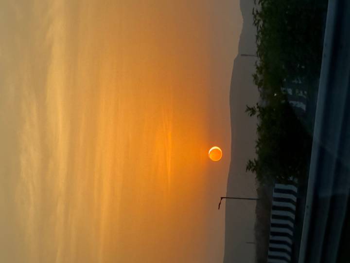Orange sunset with the sun partly eclipsed above distant hills and roadside railing silhouette.