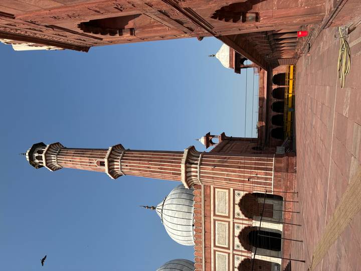 Tall red sandstone minaret and courtyard of Jama Masjid under clear blue sky.