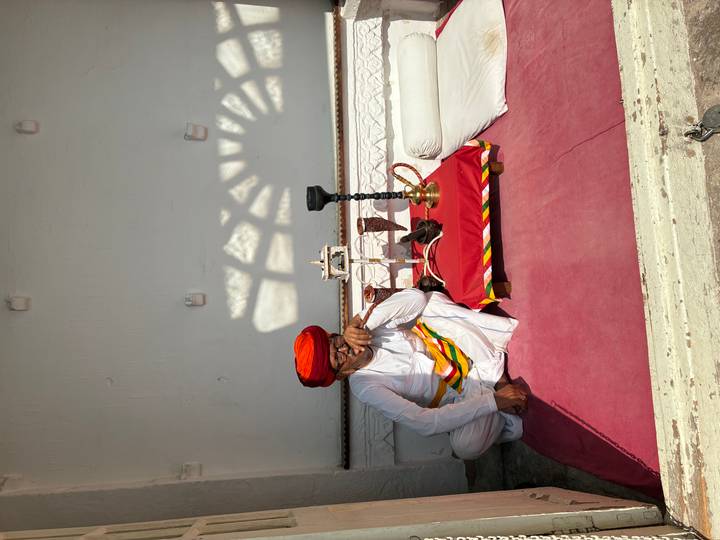 Rajasthani man in white attire and orange turban sits beside traditional hookah setup.