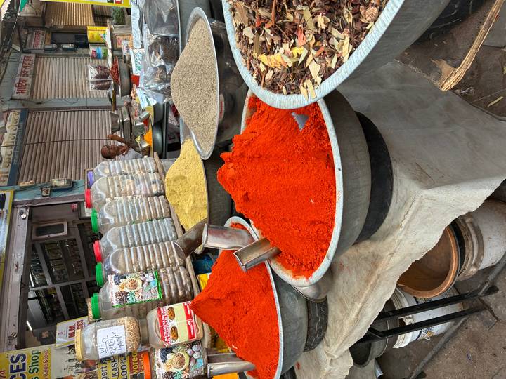 Street-side spice stall with huge metal bowls of vivid red, yellow and brown powders; a vendor sits behind the counter.