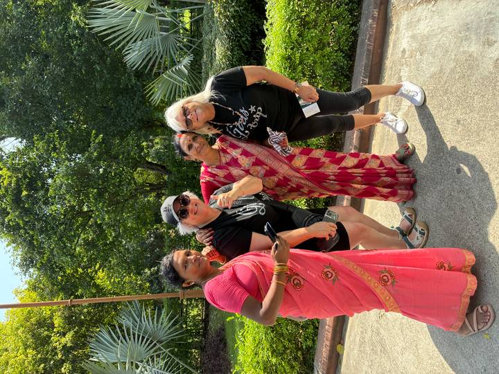 Two local women in saris and two western travellers smile together on a garden path on a bright sunny day.