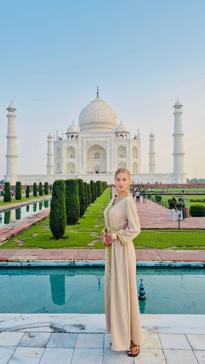 Elegant woman posing along the reflecting pool with the Taj Mahal towering behind her at dusk.