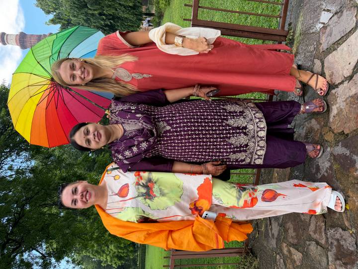 Three women smiling beneath a colorful umbrella in a garden setting with a historic stone tower rising in the background.