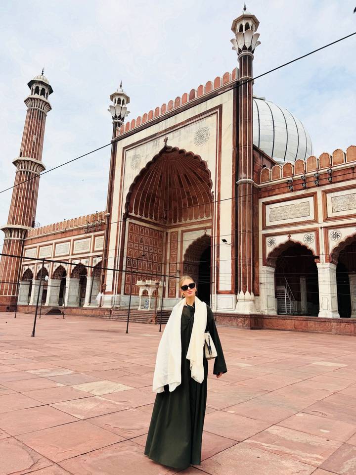 A woman stands in front of the grand arched entrance of Jama Masjid with intricate red-and-white stonework.