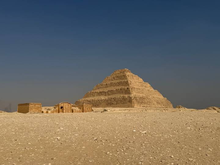 The Step Pyramid of Saqqara stands against a stark, hazy sky with small ruins at its base.