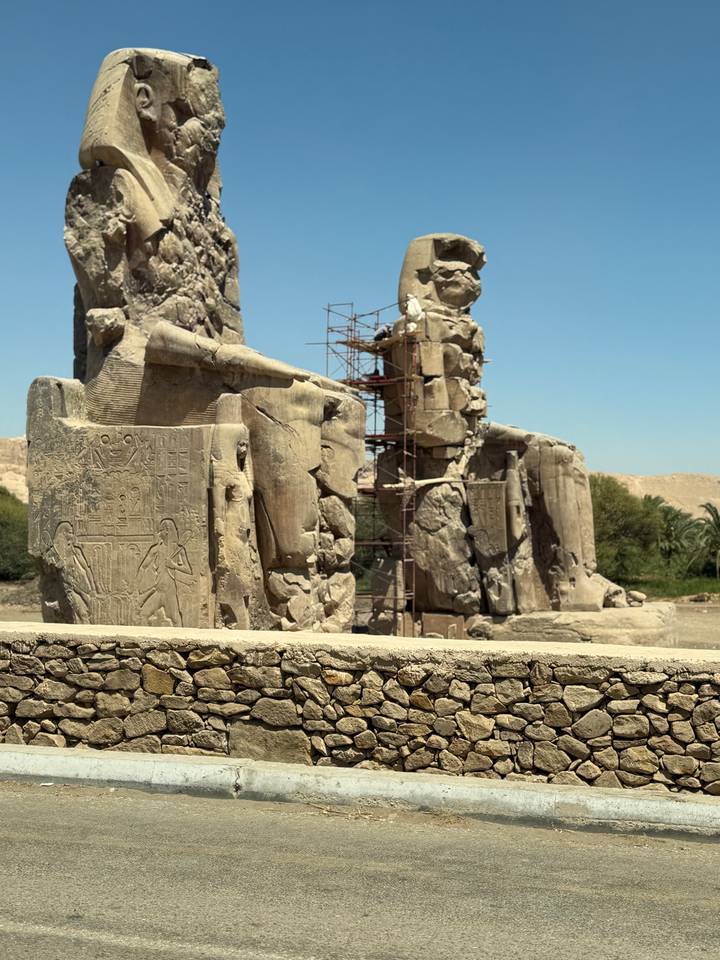 Weathered statues of the Colossi of Memnon with scaffolding amid arid surroundings.