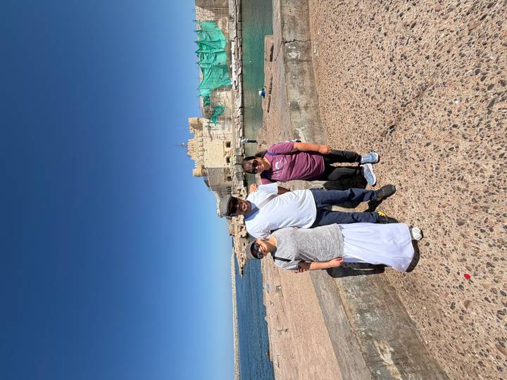 Three travelers pose beside a seaside fortress under a vivid blue sky.