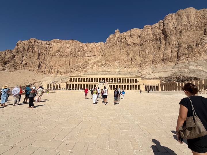 Wide courtyard view of Hatshepsut Temple with visitors walking toward its grand colonnades beneath towering cliffs.