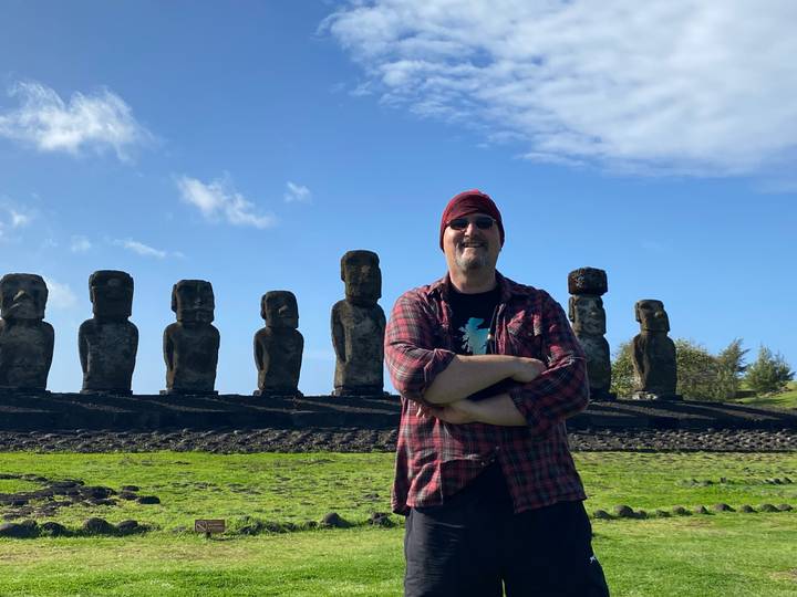 Traveler smiles with arms folded in front of a row of moai under a bright blue sky.