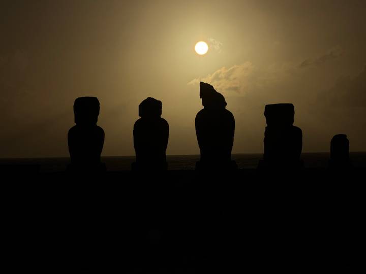 Moai silhouettes form a dark profile against a dramatic sunset with the sun low over the Pacific.