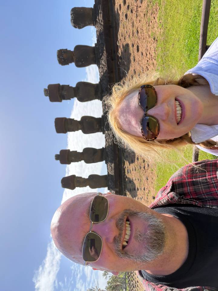 Partial selfie of a couple with a distant row of moai statues under bright daylight.