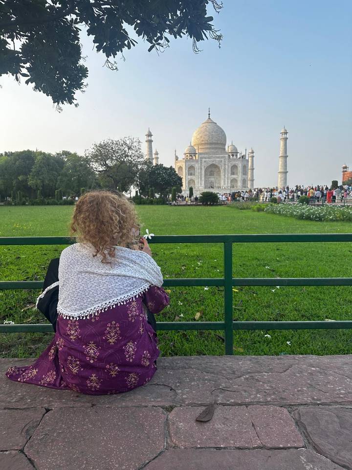 A child wrapped in a shawl gazes at the Taj Mahal from behind a green railing across a lush lawn.
