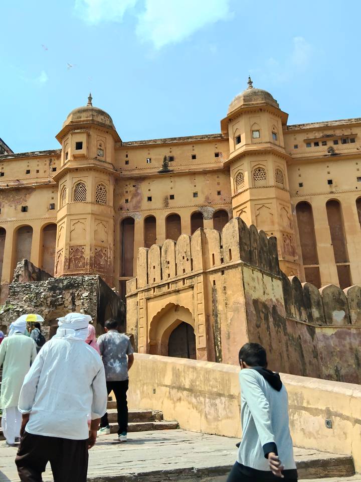 Visitors with umbrellas approach the massive yellow walls and gate of Amber Fort.