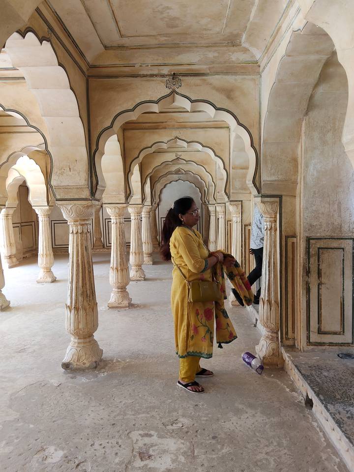 Woman in yellow saree admires a hall of carved sandstone columns and arches.