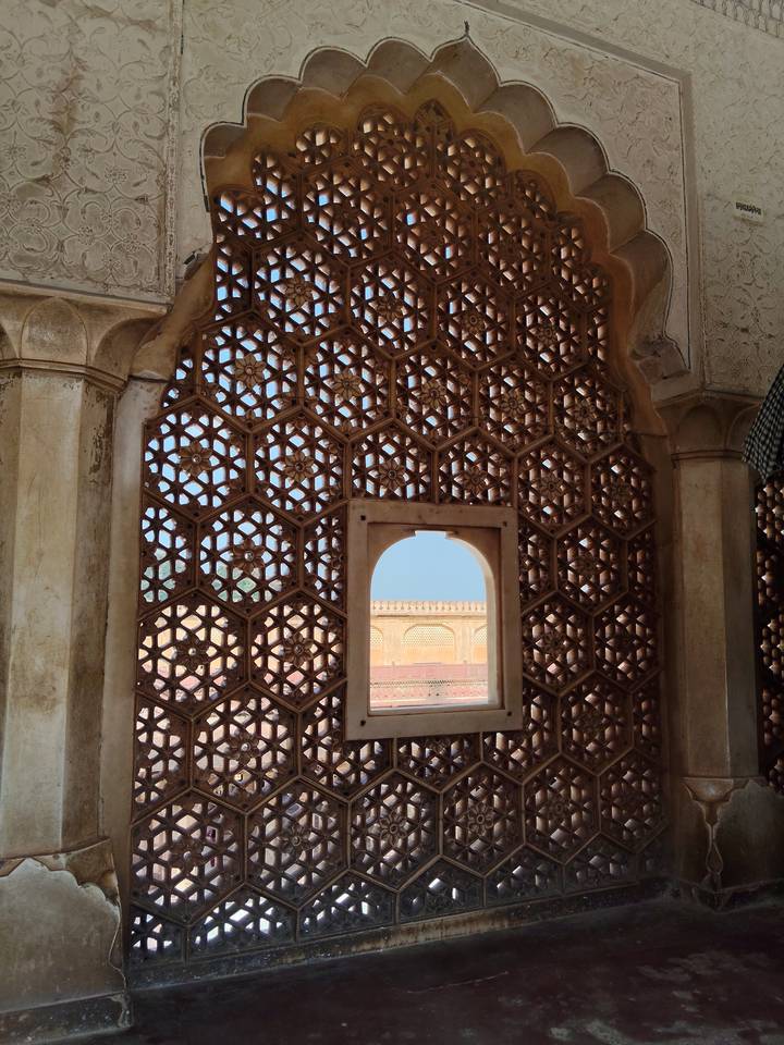 Hexagonal lattice window with a small arched opening revealing a sunny courtyard outside.