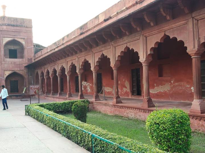 Long red-stone arcade with scalloped arches inside a Mughal complex with a lone visitor.